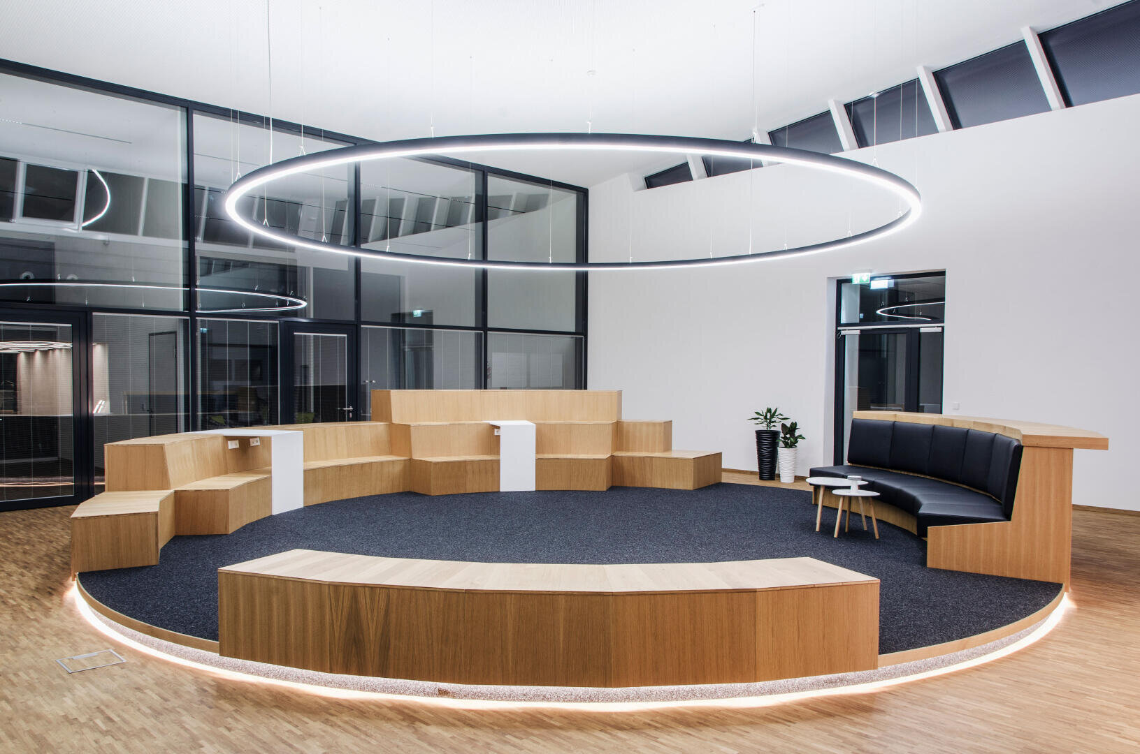 Ein moderner, runder Besprechungsraum mit abgestuften Holzsitzen, einer schwarzen, gepolsterten Bank, kleinen Tischen und einer großen, runden Deckenleuchte in einem Raum mit Glaswänden und Holzböden.