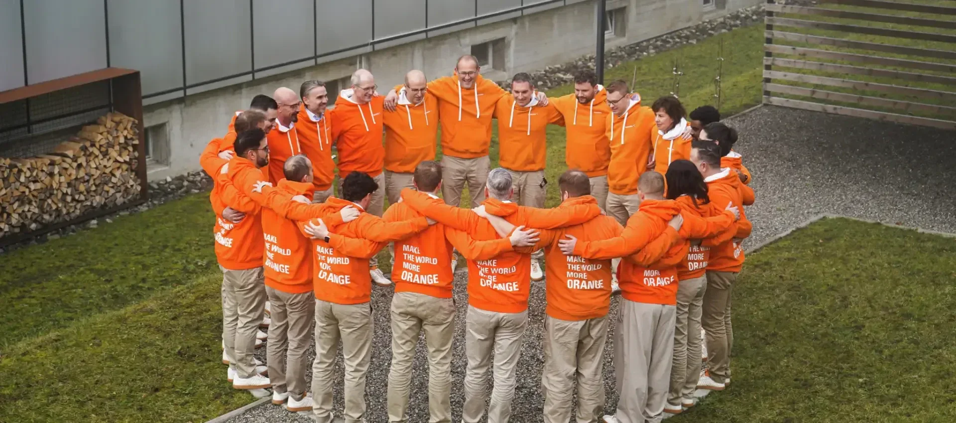 Un groupe de personnes vêtues de sweats à capuche orange et de pantalons beiges se tient en cercle à l'extérieur, les bras croisés, sur une pelouse à proximité d'un bâtiment.