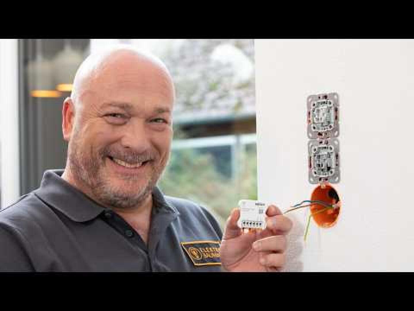 Ein lächelnder Mann in einem grauen Polohemd hält ein elektrisches Gerät neben einer offenen Wandsteckdose mit freiliegenden Drähten und scheint dabei zu sein, es zu installieren oder zu reparieren.