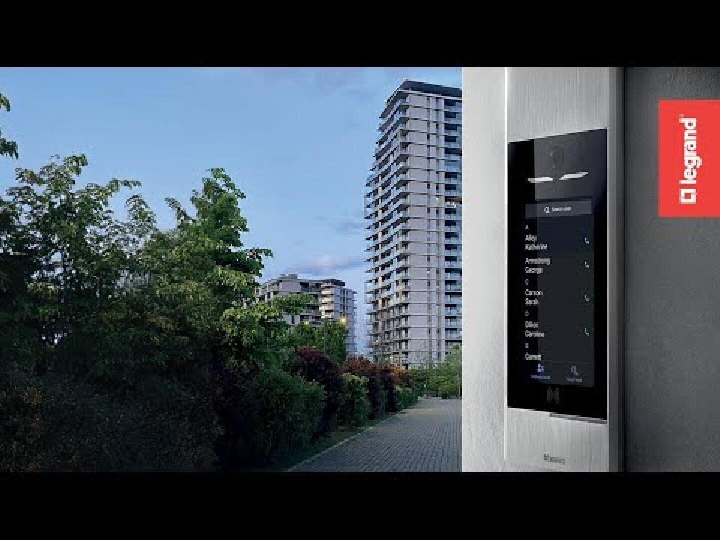 Un immeuble résidentiel moderne sous un ciel bleu avec un interphone vidéo intelligent monté sur un mur au premier plan ; le logo Legrand apparaît sur le côté droit de l'image.