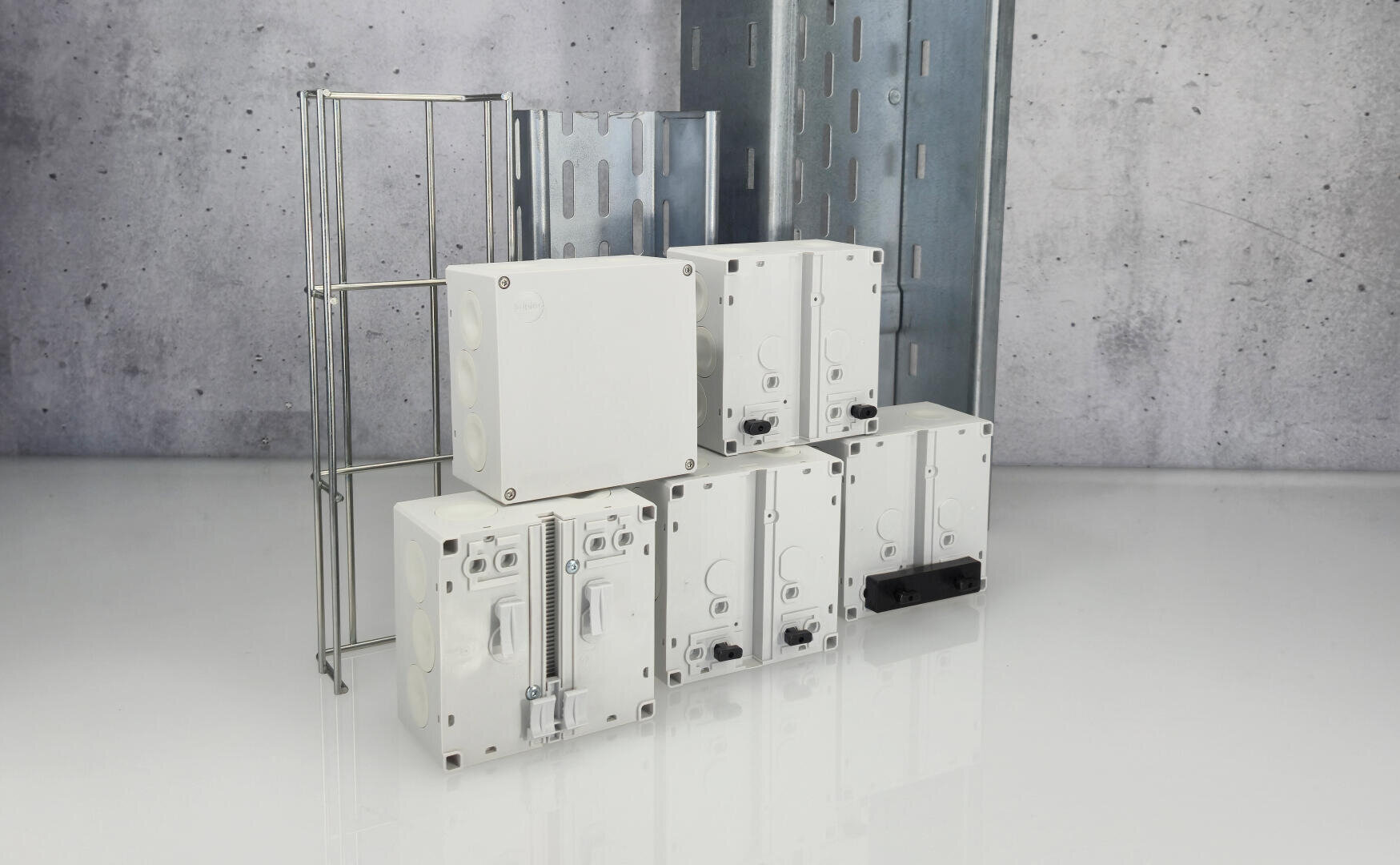 Gruppe von weißen elektrischen Abzweigdosen und Metallzubehör für das Kabelmanagement, die auf einer reflektierenden Oberfläche angeordnet sind, mit einem Hintergrund aus grauem Beton im Industriestil.