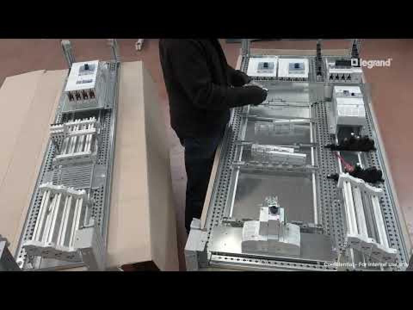 Eine Person, die elektrische Schalttafeln mit verschiedenen Bauteilen und Verkabelungen innerhalb eines Metallrahmens zusammenbaut oder inspiziert, von oben gesehen. Das Legrand-Logo ist in der oberen rechten Ecke zu sehen.