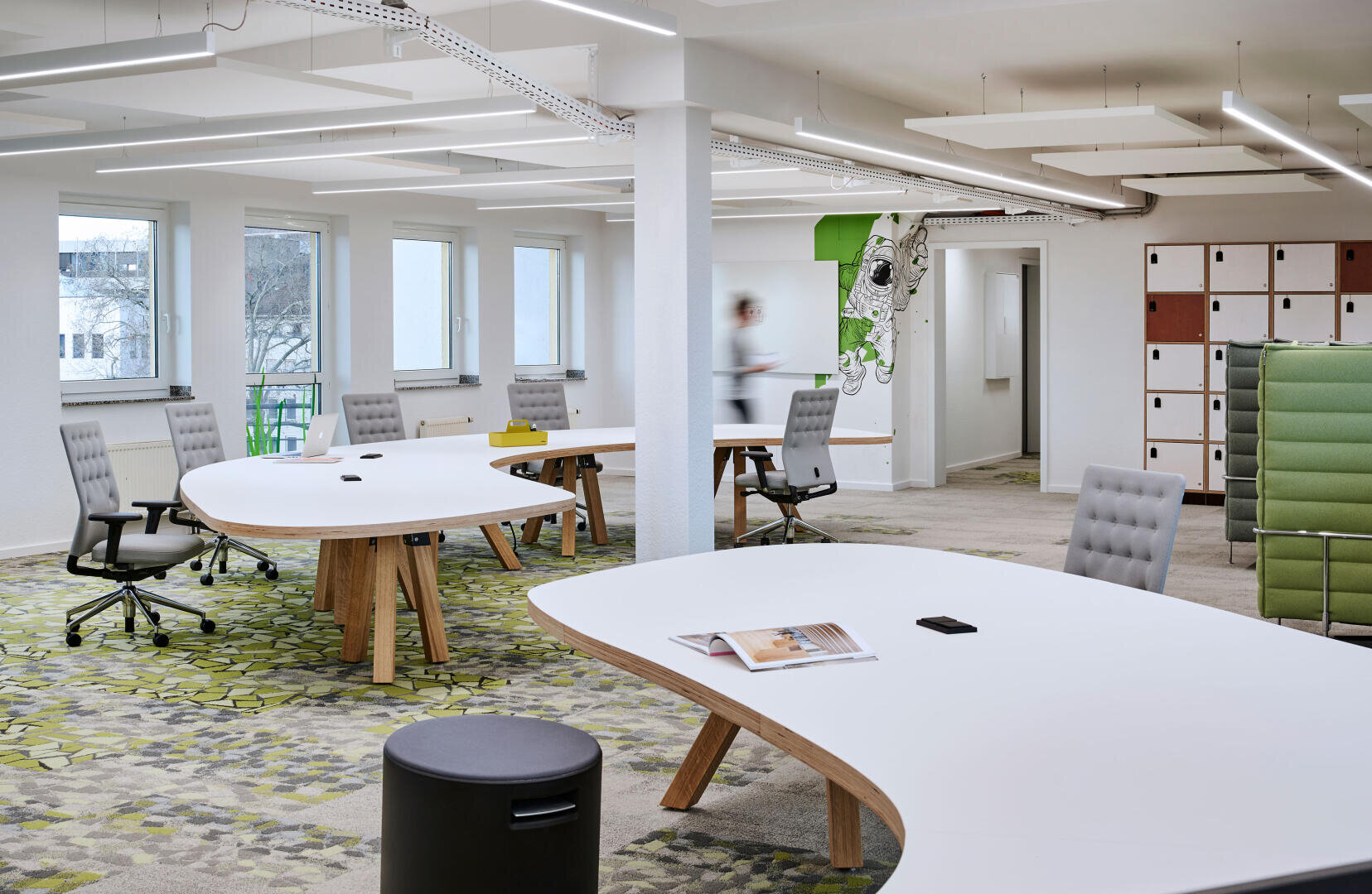 Modernes Großraumbüro mit großen, gebogenen weißen Schreibtischen, ergonomischen Stühlen und gemustertem Teppichboden. Schließfächer, Fenster und eine verschwommene Person, die im Hintergrund läuft, schaffen einen sauberen und luftigen Arbeitsbereich.
