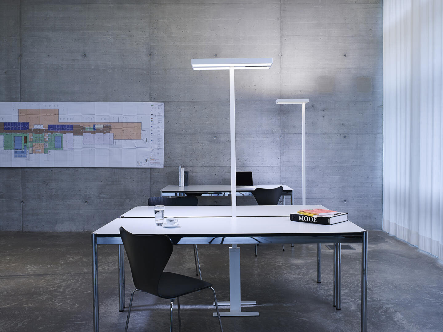 Ein modernes, minimalistisches Büro mit zwei Schreibtischen, schwarzen Stühlen, hohen weißen Lampen, einem Glas Wasser, einem Stapel Aktenordner und einem Buch mit dem Titel MODE vor einer Betonwand mit einem großen Architekturplan.