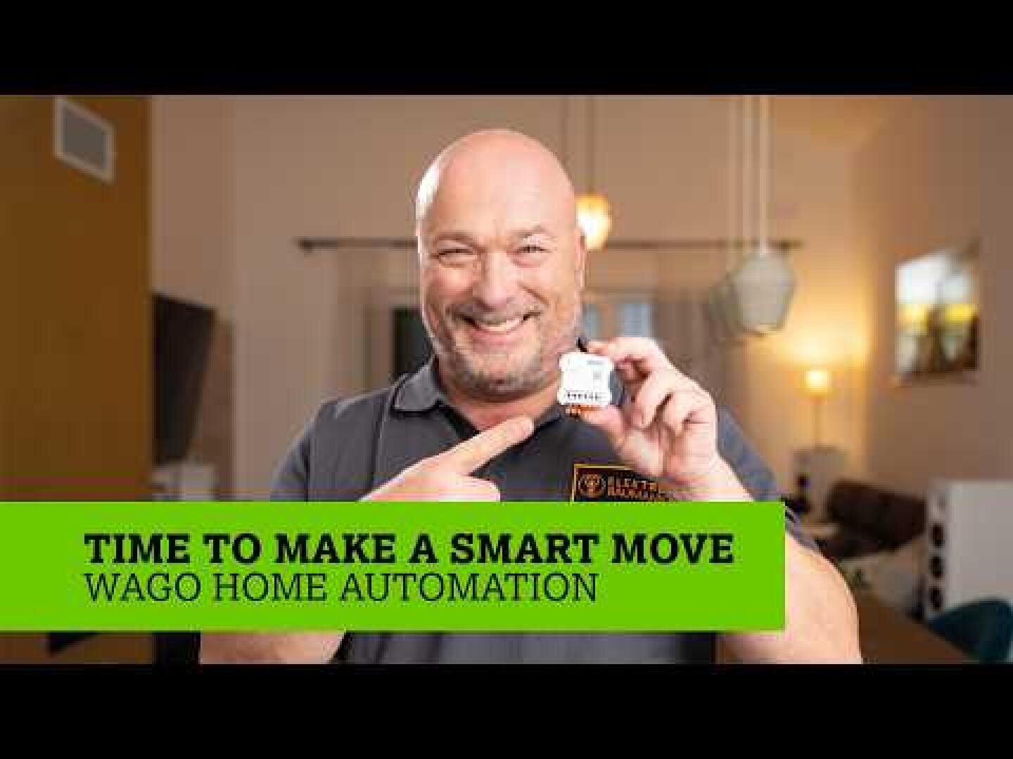 Dans un salon lumineux, un homme souriant montre du doigt un petit appareil qu'il tient à la main. Une bannière verte en bas de l'écran indique : "Il est temps de faire un geste intelligent. Wago Home Automation.