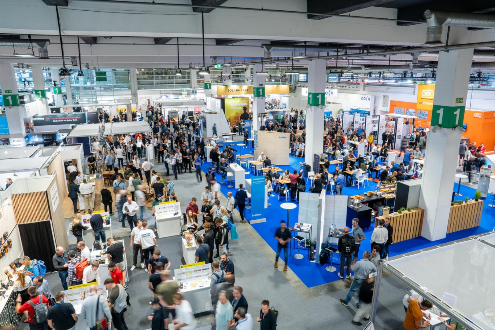 Un grand hall de congrès très animé dans lequel de nombreuses personnes se promènent et visitent différents stands. Des tapis bleus bordent les allées et les exposants présentent des produits et des bannières sur leurs stands. L'espace est brillamment éclairé et animé.