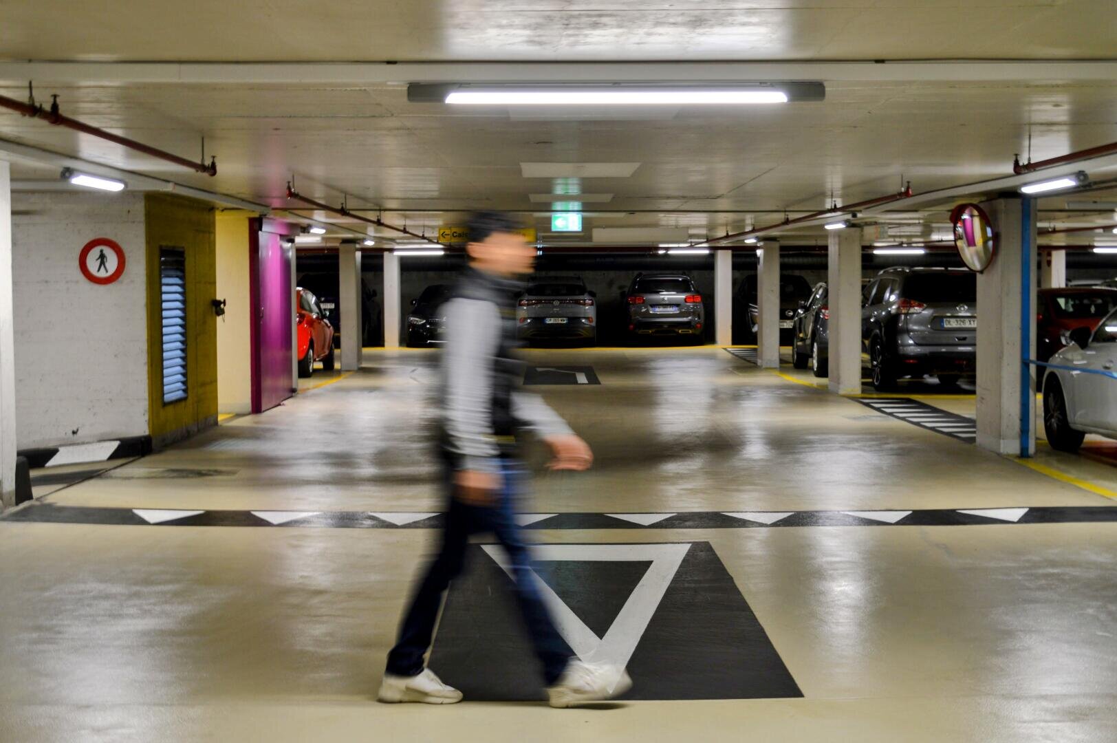Une personne en mouvement traverse un parking souterrain avec des voitures garées des deux côtés et un éclairage fluorescent. L'image est légèrement floue, ce qui accentue le mouvement.