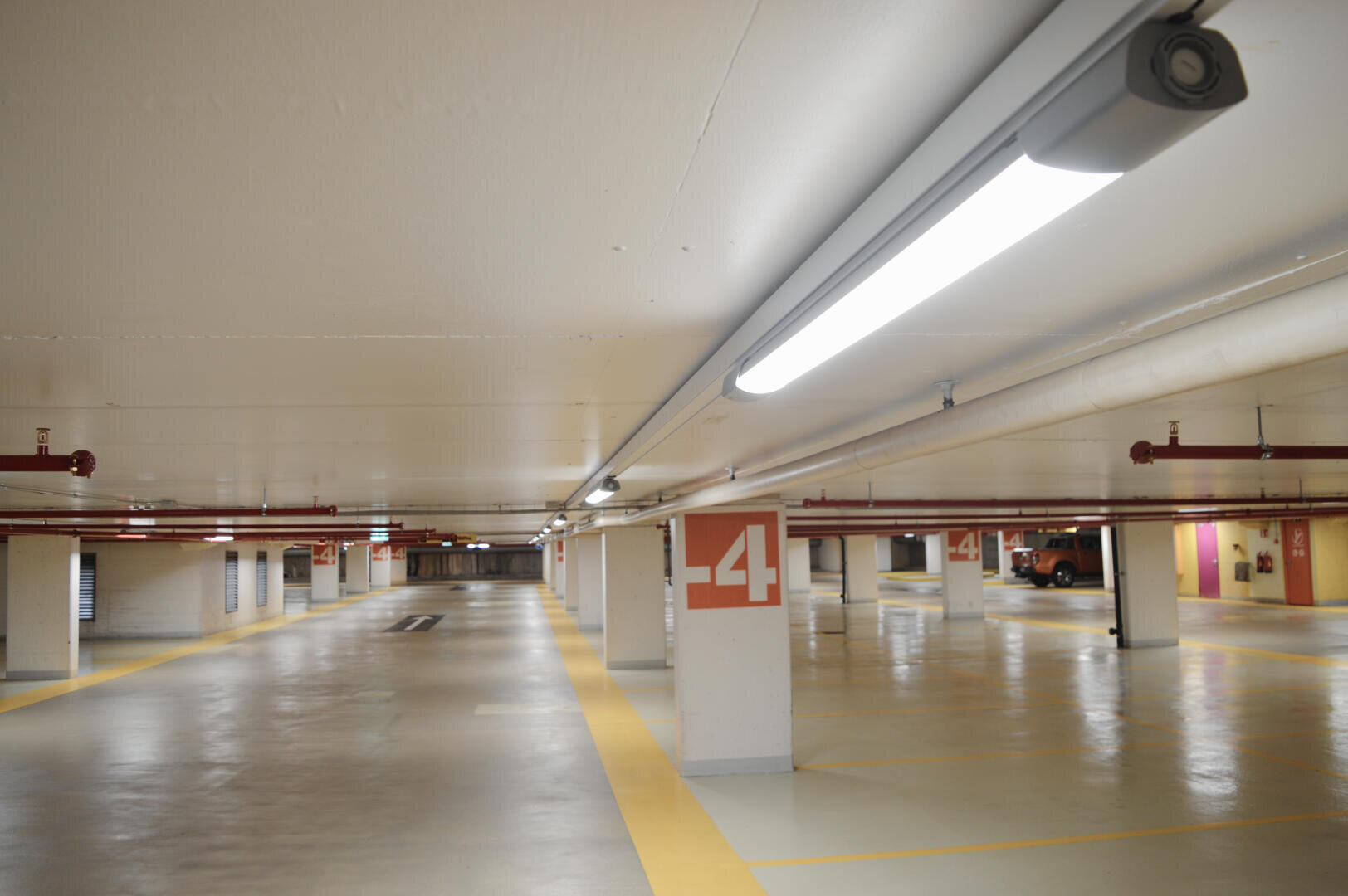 Un parking intérieur vide et bien éclairé avec des murs blancs, des lignes jaunes et des colonnes marquées "-4". Quelques voitures garées sont visibles à l'arrière-plan.
