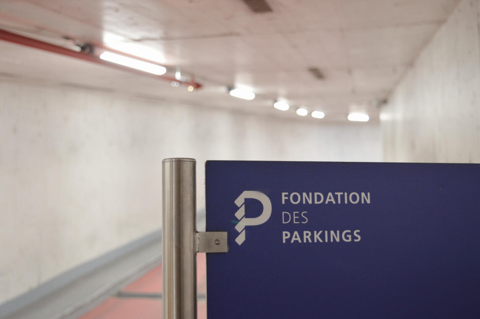 Un panneau bleu indiquant Fondation des Parkings en français est monté sur un poteau métallique à l'intérieur d'un parking souterrain ou d'un tunnel aux murs blancs et bien éclairés.