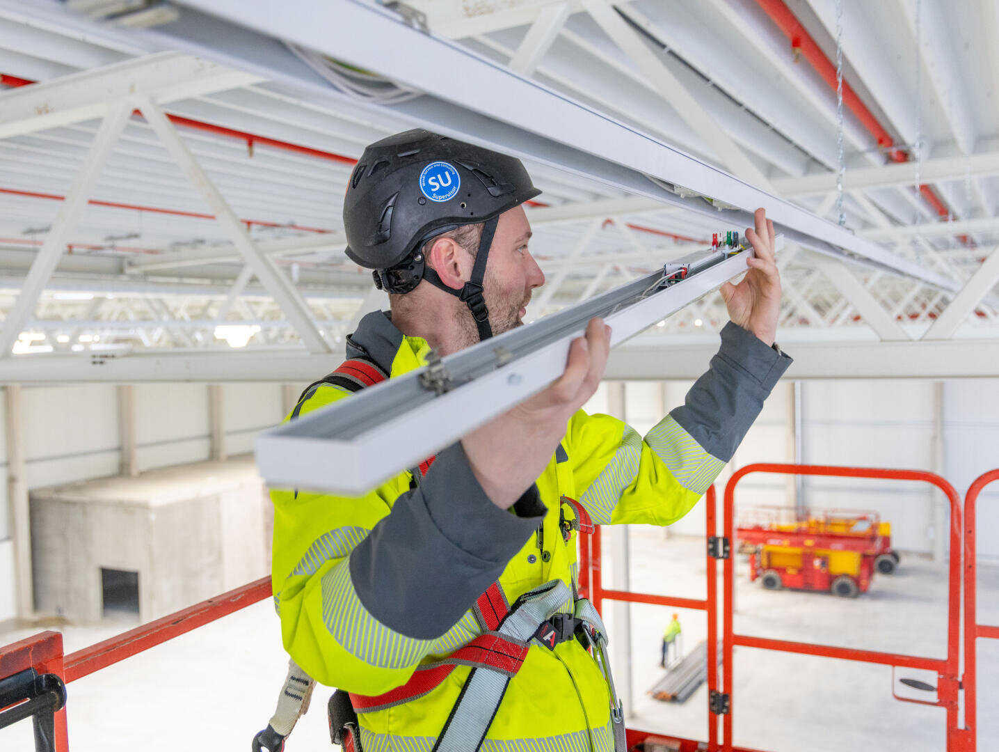 Un ouvrier du bâtiment portant un casque et un gilet de sécurité monte un plafonnier alors qu'il se trouve sur un ascenseur dans un grand bâtiment industriel.