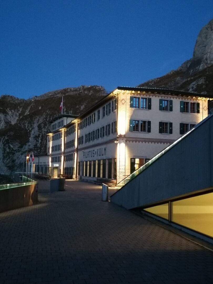 Ein grosses, beleuchtetes Gebäude mit der Aufschrift "Pilatus-Kulm" steht in der Abenddämmerung vor einer Bergkulisse mit einem gepflasterten Gehweg und modernen Glasgeländern im Vordergrund.