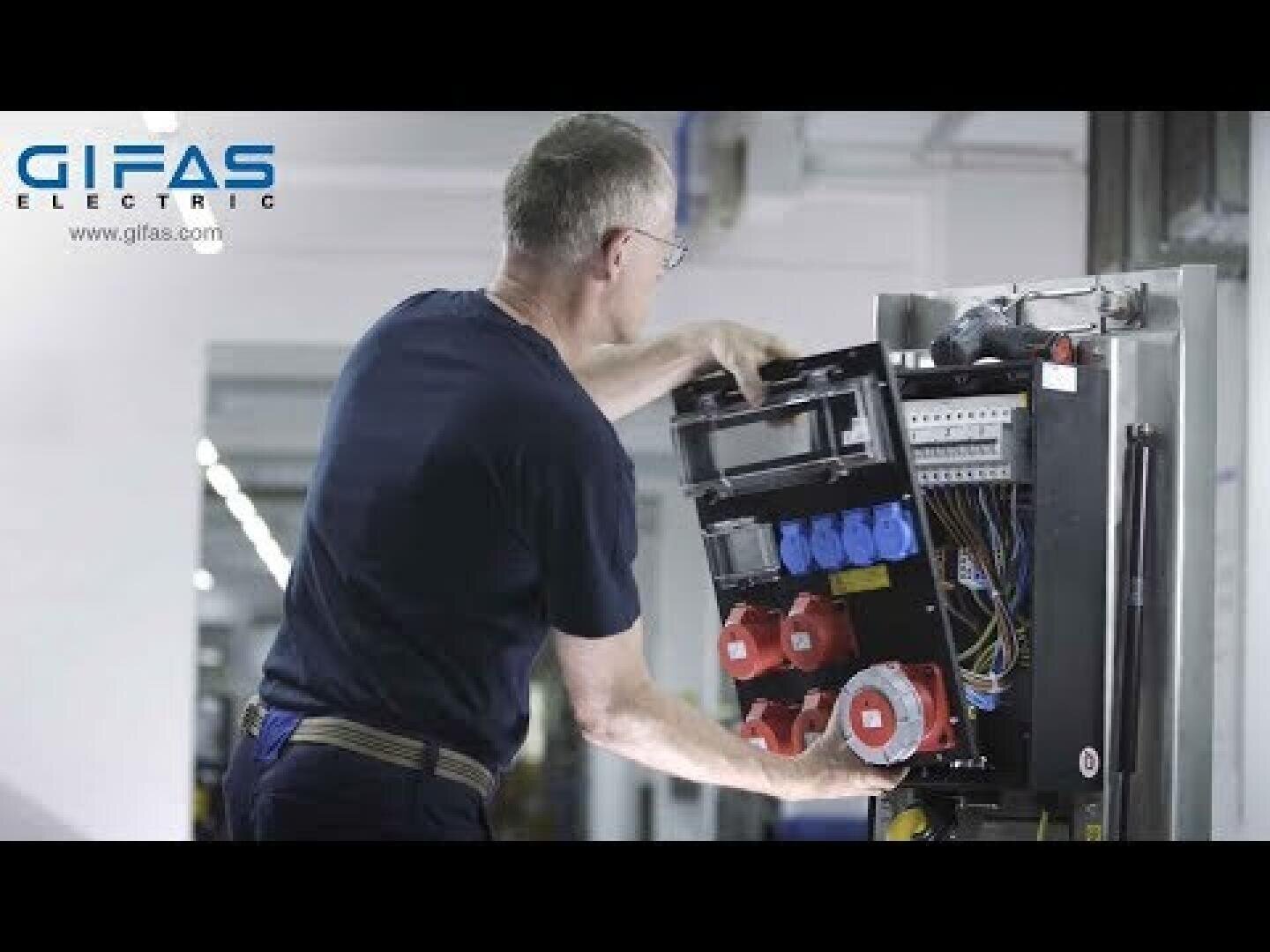 Ein Mann mit Brille arbeitet an einer offenen elektrischen Schalttafel mit mehreren farbigen Steckdosen und Kabeln. Das Logo der GIFAS Electric ist in der oberen linken Ecke zu sehen.