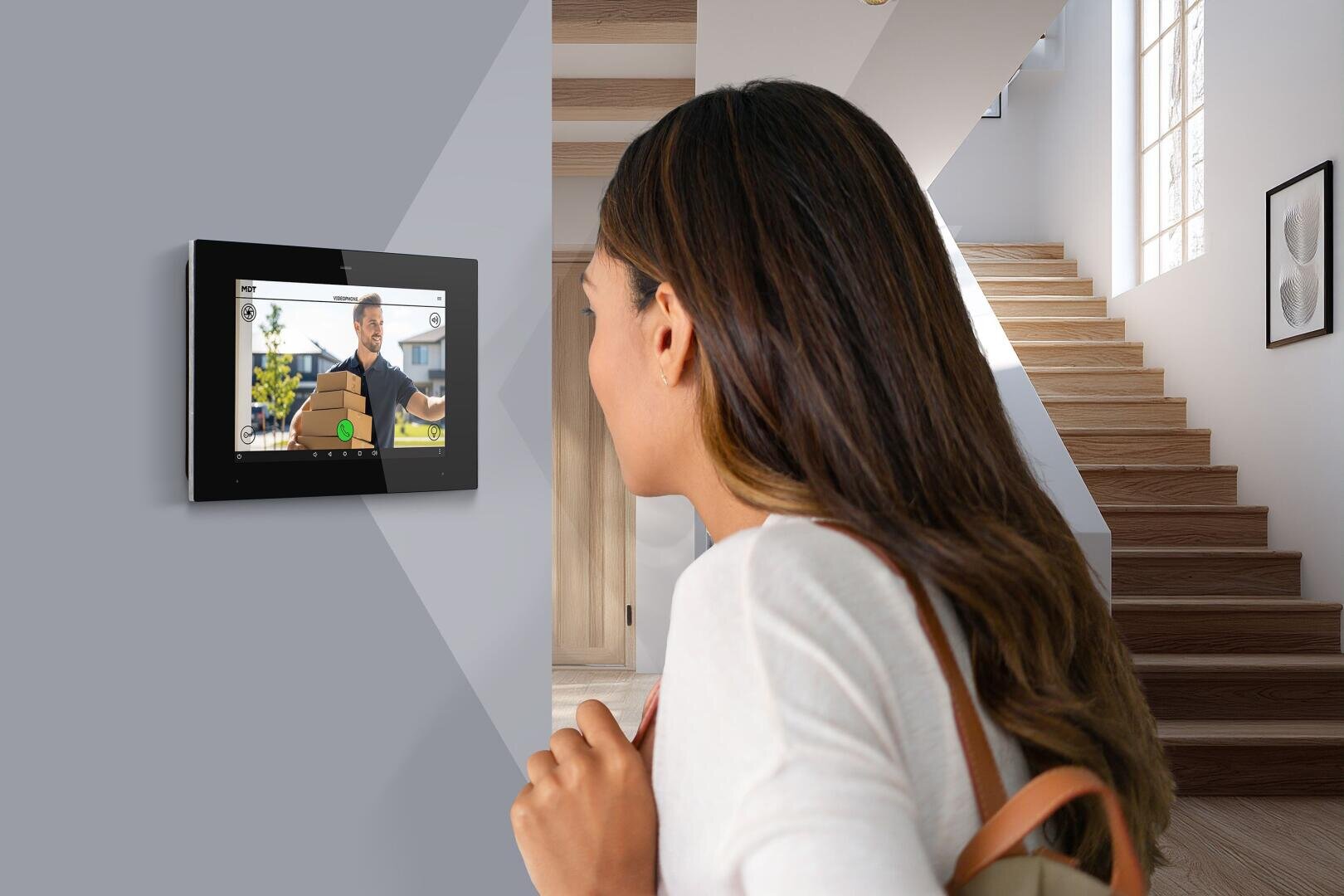 Une femme aux longs cheveux bruns se tient dans une maison et regarde un écran fixé au mur, sur lequel un homme parle par appel vidéo. Dans l'arrière-plan moderne et lumineux, on voit un escalier et une grande fenêtre.
