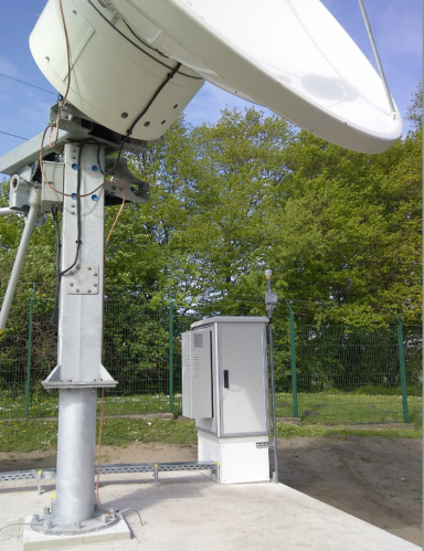 Eine große Satellitenschüssel auf einem Metallständer steht im Freien in der Nähe eines eingezäunten Geländes mit grünen Bäumen und einem grauen Geräteschrank darunter. Der Himmel ist klar und blau.