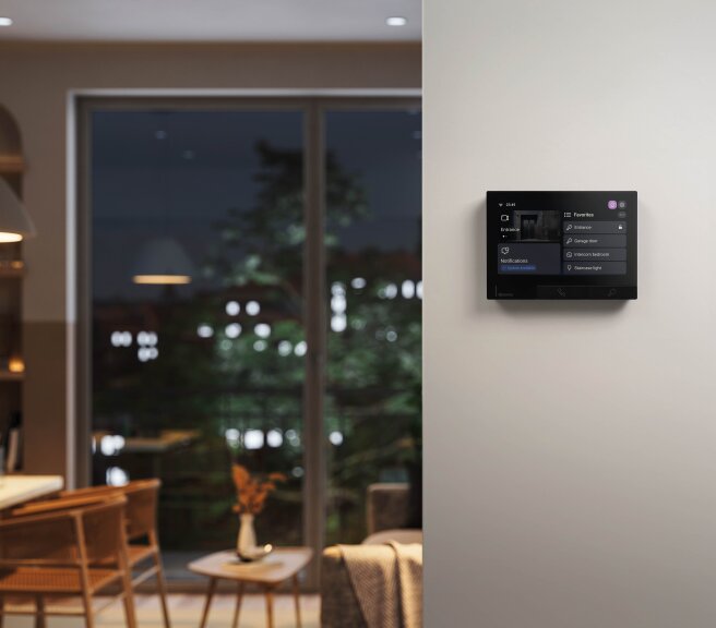 Ein Smart-Home-Bedienfeld ist in einem modernen Wohnzimmer an der Wand montiert, mit großen Fenstern, auf denen Bäume im Freien zu sehen sind, und Umgebungslicht im Hintergrund.