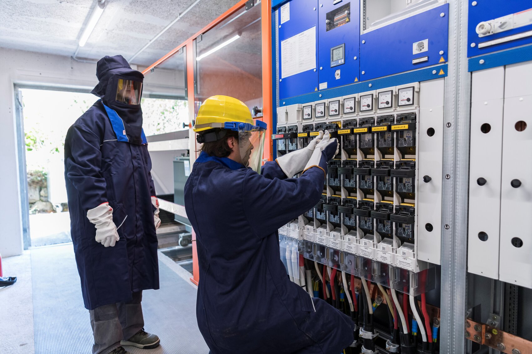 Deux ouvriers portant des vêtements de protection et un casque vérifient et actionnent des interrupteurs électriques sur un grand tableau électrique dans une installation industrielle. Un ouvrier est à genoux et règle les interrupteurs, tandis que l'autre se tient à proximité.